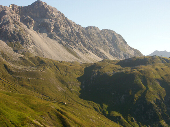 Grüne Berghänge unterhalb schroffer Felsgipfel bei St. Anton am Arlberg, durchzogen von Wanderwegen und Almflächen im Spätsommer.