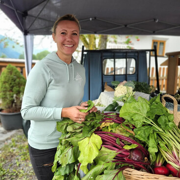 Miriam Scherl mit blondem Dutt steht hinter einem Marktstand. Vor ihr liegen frischer Mangold, Rüben und anderes regionales Gemüse. Sie lächelt freundlich in die Kamera.