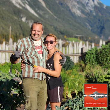Miriam und Dave stehen lachend in einem Gemüsebeet mit Bergen im Hintergrund. Dave  trägt Arbeitshandschuhe und hält eine kleine gelbe Rübe, Miriam  trägt eine Sonnenbrille. Beide umarmen sich.