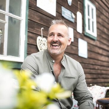 David Bradley mit grauem Haar und Dreitagebart sitzt lächelnd vor einer rustikalen Holzwand mit Fenster. Im Vordergrund verschwommene Blumen.