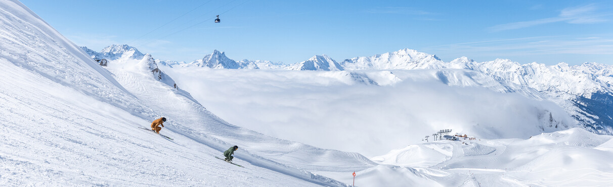 Two skiers descend a snowy slope in sunshine with panoramic alpine views.