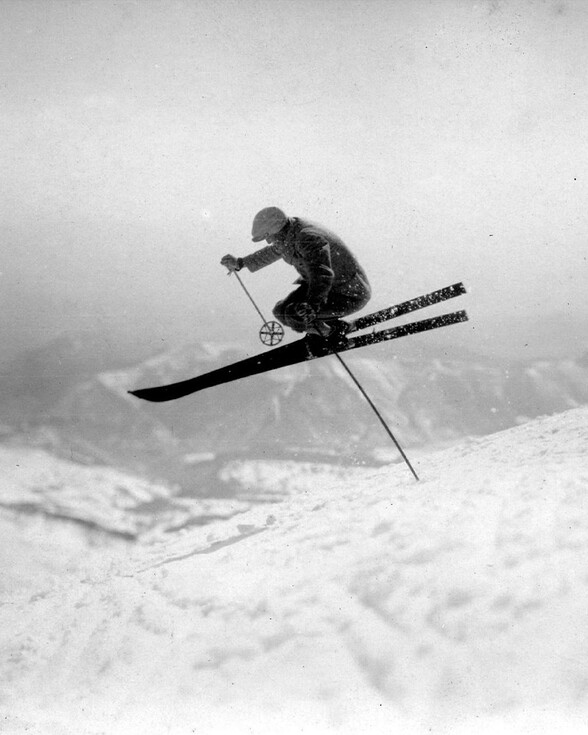 Historische Schwarzweißaufnahme von Hannes Schneider in der Luft – dynamischer Sprung in tiefer Hocke über eine verschneite Piste in den Alpen bei St. Anton am Arlberg.