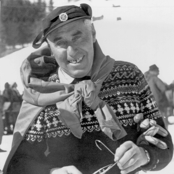 Schwarzweißporträt von Hannes Schneider in traditionellem Norwegerpullover und Barett, lächelnd vor winterlicher Kulisse in St. Anton am Arlberg.
