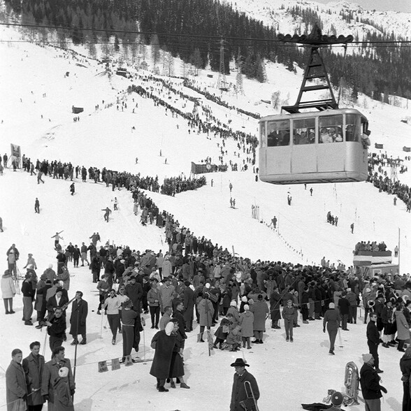 Schwarzweißaufnahme eines historischen Wintersportevents in St. Anton am Arlberg: Menschenmengen säumen die Skipiste, während eine Seilbahnkabine über das Geschehen schwebt.