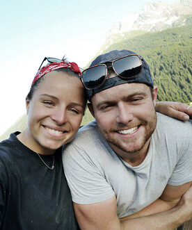 Ein junges Paar lächelt fröhlich in die Kamera vor einer Berglandschaft bei klarem Himmel. Beide tragen Freizeitkleidung, die Frau ein rotes Bandana.