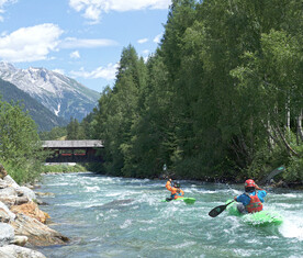 Zwei Kajakfahrer paddeln einen reißenden Gebirgsfluss entlang. Im Hintergrund sind eine überdachte Holzbrücke und die Alpen sichtbar.