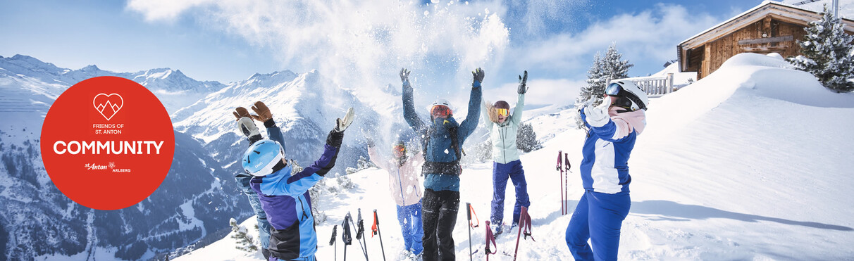 Skifahrer in bunter Winterkleidung jubeln im Schnee vor Alpenpanorama und Hütte. Kreisförmiges "Friends of St. Anton – Community"-Logo links im Bild.