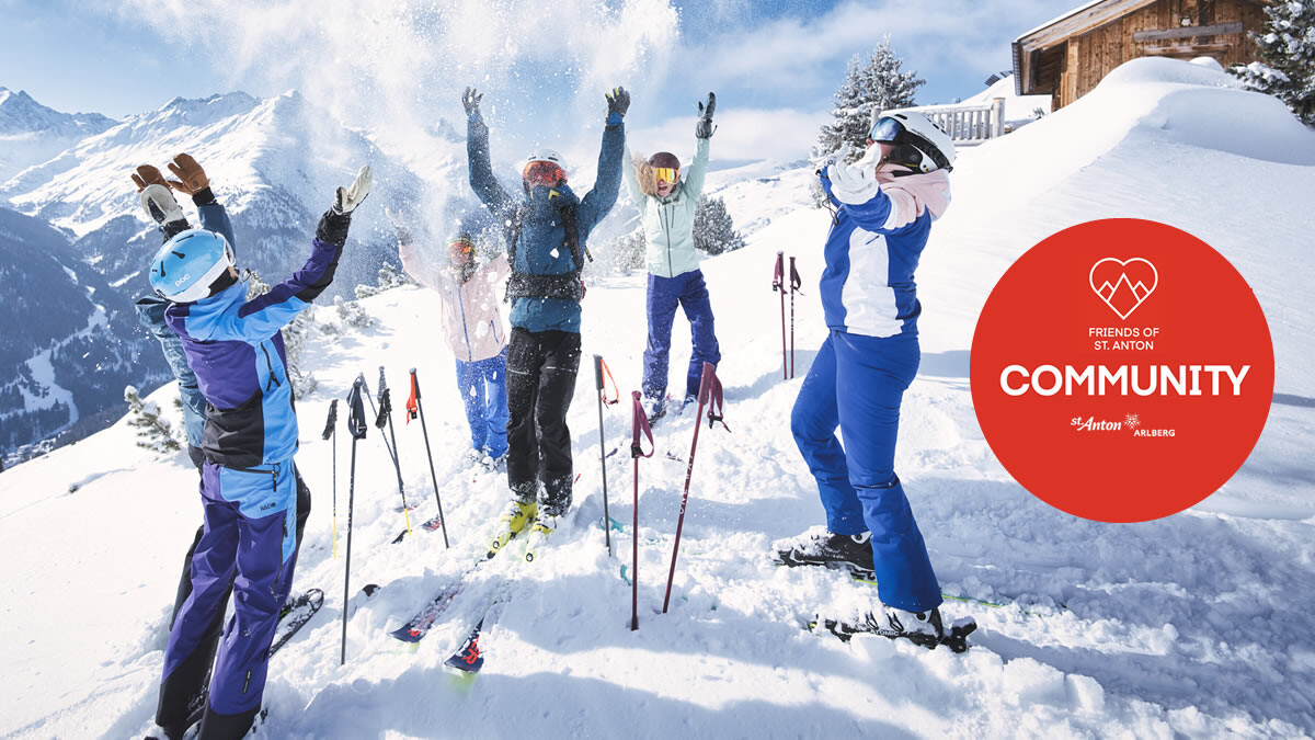 A joyful ski group stands in front of a snow-covered alpine hut in St. Anton am Arlberg, throwing snow into the air together. A red circle in the foreground features the “Friends of St. Anton” logo and the word “COMMUNITY.” Snowy mountain peaks stretch into the distance under a blue sky. The image conveys a sense of community and winter fun in the Tyrolean Alps.