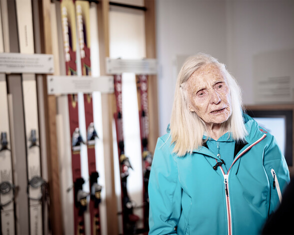 Eine ältere Frau mit hellblauer Jacke steht vor historischen Skiern in einem Museum. Sie schaut ernst und konzentriert.