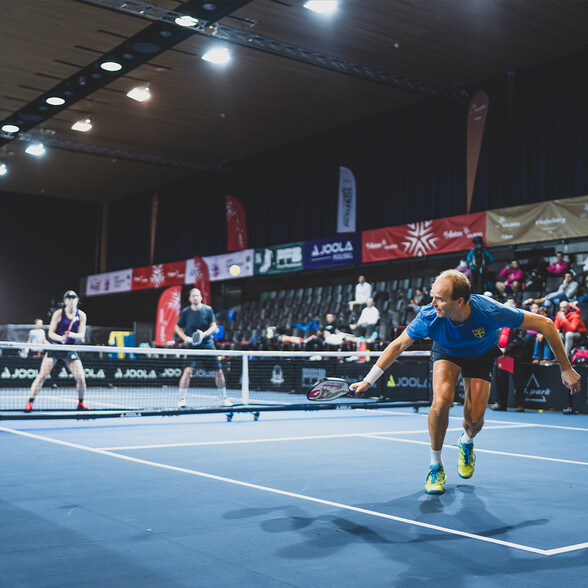 n einer modernen Sporthalle spielt ein Mixed-Doppel Pickleball. Im Vordergrund ist ein Spieler beim schnellen Vorstoß zu sehen.
