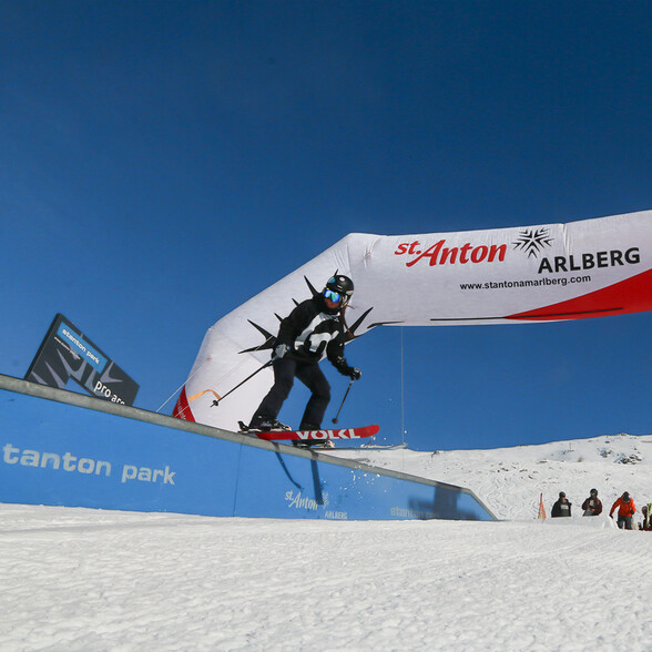Freestyle-Skifahrer auf einem Rail im Stanton Park, vor blauem Himmel und mit St. Anton-Branding im Hintergrund. Wintersport-Action pur.