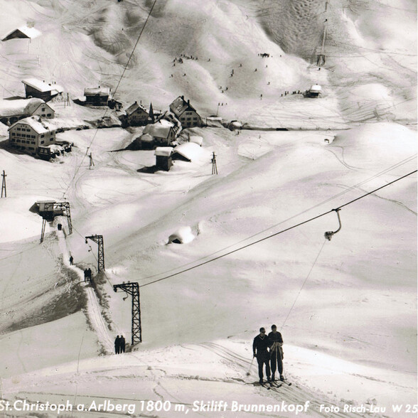 Historische Schwarzweißaufnahme eines Skigebiets mit Schlepplift in St. Christoph am Arlberg. Mehrere Skifahrer sind zu sehen.