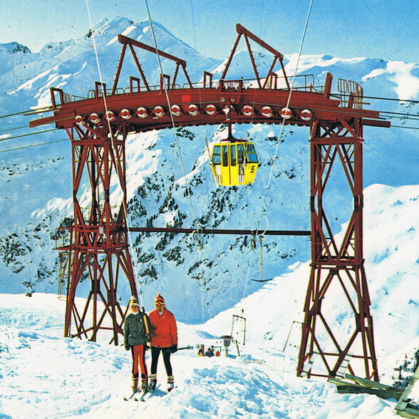 Eine gelbe Seilbahngondel hängt an einer roten Stahlkonstruktion in den verschneiten Alpen. Zwei Skifahrer stehen unter der Gondel.