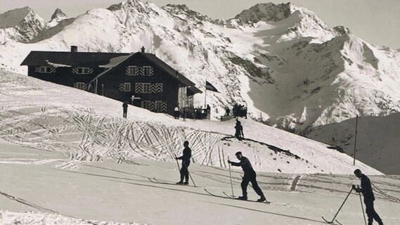 Three skiers standing on a mountian platue in fresh powder snow, leaving visible tracks behind them.