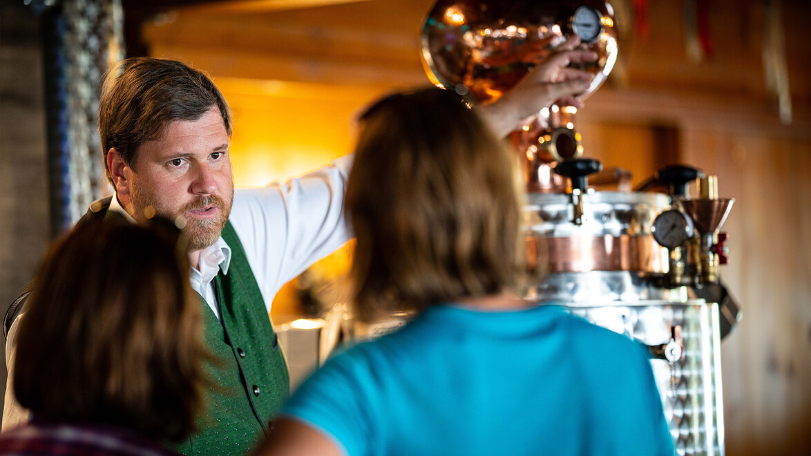 Mann erklärt Brennerei-Prozess an Kupferbrennkessel, Gäste hören aufmerksam zu / Man explains distillation process using a copper still, guests listen attentively