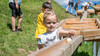 Kind spielt mit Holzkugelbahn im Freien, weitere Kinder im Hintergrund / Child plays with a wooden marble run outdoors, other children in the background