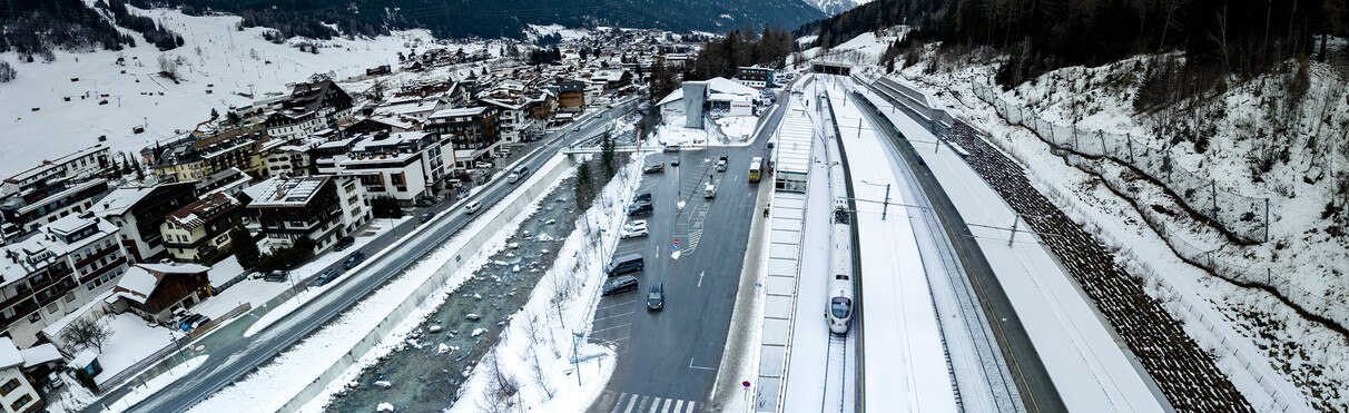 Luftaufnahme des Bahnhofs St. Anton mit einem ICE-Zug auf verschneiten Gleisen und dem Ort im Hintergrund. Umweltfreundliche Anreise sichtbar gemacht.