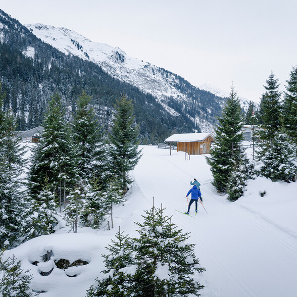 Two cross-country skiers glide along a groomed trail through the snowy landscape near St. Anton am Arlberg. They are surrounded by snow-covered trees, wooden huts, and majestic alpine peaks.
