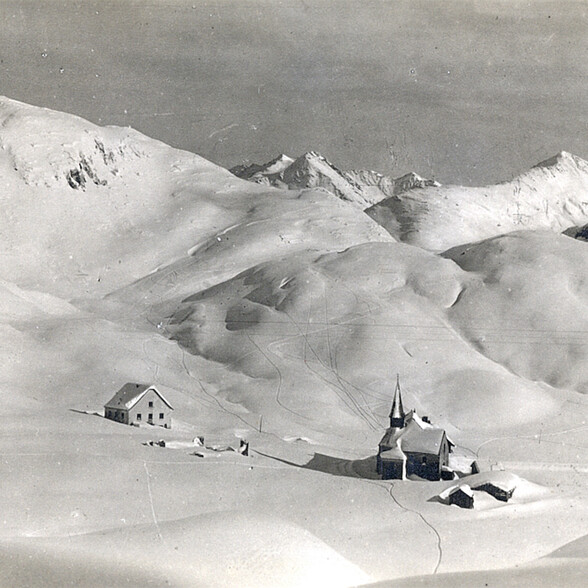 Historische Winteraufnahme von St. Christoph am Arlberg mit tief verschneiter Kapelle inmitten der Alpen. Die unberührte Schneelandschaft und die stille Atmosphäre zeigen den Ort, wie er früher war.