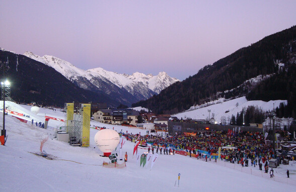 Abendliches Bild eines Skirennens mit Menschenmenge am Zielgelände von St. Anton am Arlberg. Die Alpen im Hintergrund färben sich im Licht der untergehenden Sonne.