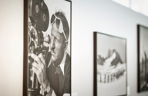 Porträtfoto von Stefan Kruckenhauser, Pionier des alpinen Skilaufs, wie er durch eine alte Filmkamera blickt. Das Bild erinnert an die frühen Tage des Skifilms in St. Anton am Arlberg.