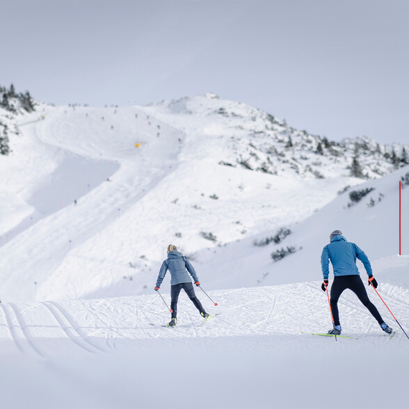 Zwei Langläufer gleiten im klassischen Stil auf einer Loipe durch tief verschneites, offenes Gebirgsgelände mit Aussicht auf Skispuren.
