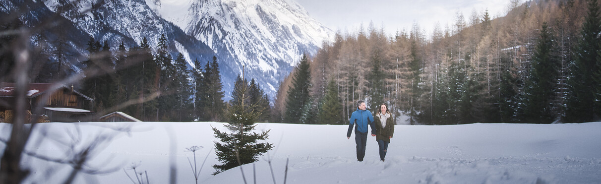 Couple walking through a snowy valley with mountain and forest backdrop.