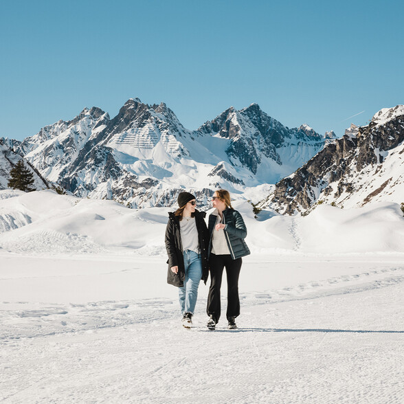 Two women walking on a cleared winter path with snowy mountains behind.
