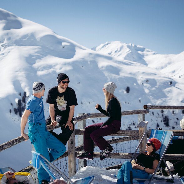 Vier Freunde genießen entspannte Momente auf einer sonnigen Winterterrasse in den Alpen, umgeben von tief verschneiten Berghängen.