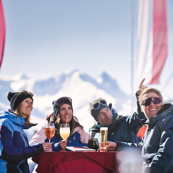 Eine Gruppe Menschen genießt bunte Getränke an einem gedeckten Tisch mit Blick auf die verschneiten Gipfel von St. Anton am Arlberg.