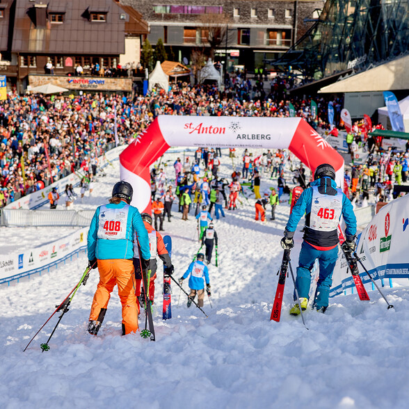 Teilnehmer des Kult-Skirennens „Der Weisse Rausch“ warten am Start unter einem großen Bogen mit der Aufschrift „St. Anton am Arlberg“, umgeben von jubelnden Zuschauern.