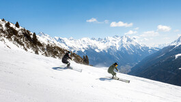 Zwei Skifahrer fahren synchron eine sonnige Piste mit Panoramablick ins Stanzertal hinab.