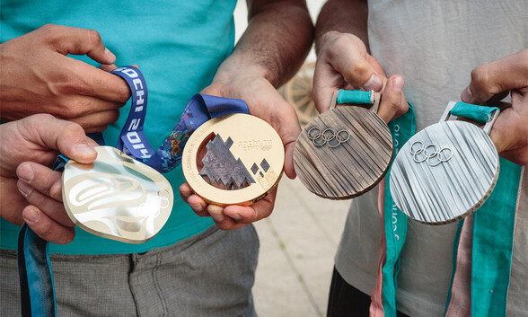 Vier Hände präsentieren stolz olympische Gold-, Silber- und Bronzemedaillen mit markanten Gravuren und farbigen Bändern.
