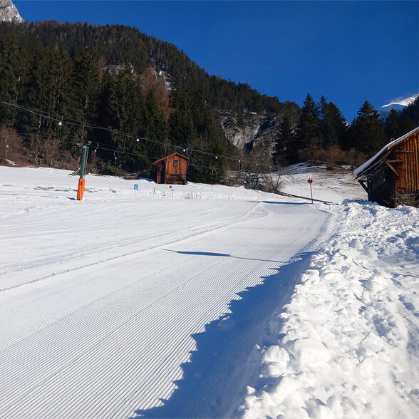 Eine frisch präparierte Skipiste mit Spurgerillen führt an alten Holzhütten vorbei zu einem nostalgischen Skilift im Waldgebiet von St. Anton.