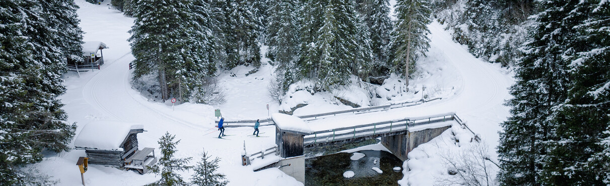 Zwei Langläufer überqueren eine verschneite Holzbrücke im Winterwald bei St. Anton.