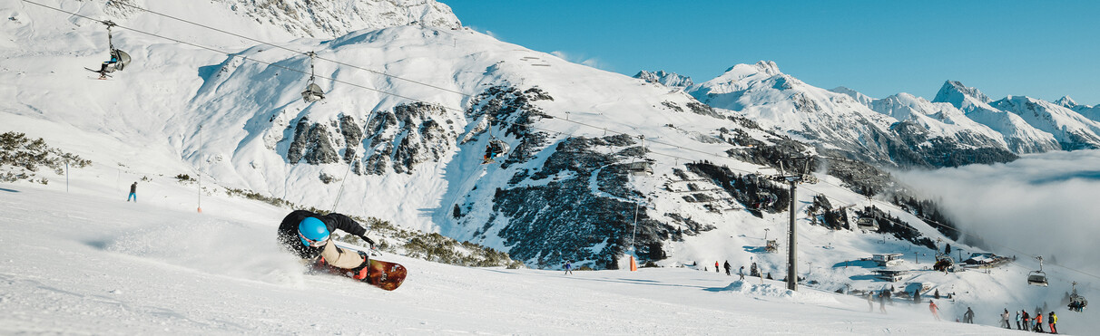 Snowboarder carves dynamically down a slope with chairlift and snowy Arlberg peaks in the background.