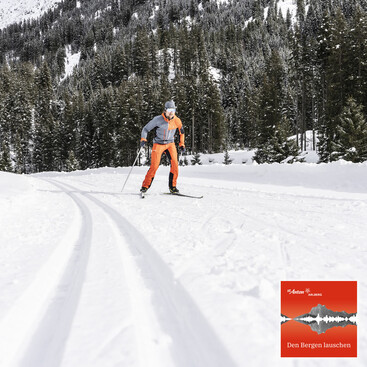 Ein Langläufer in oranger Sportkleidung fährt auf einer frisch präparierten Loipe durch eine verschneite Winterlandschaft mit dichten Tannen.