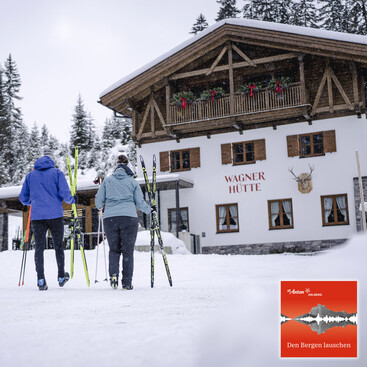 Zwei Langläufer:innen tragen ihre Ski und gehen auf eine verschneite Berghütte mit dem Namen „Wagner Hütte“ zu. Die Hütte ist weihnachtlich dekoriert.