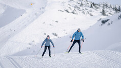 Zwei Personen beim Langlaufen auf einer sonnigen Loipe im schneebedeckten Gebirge.