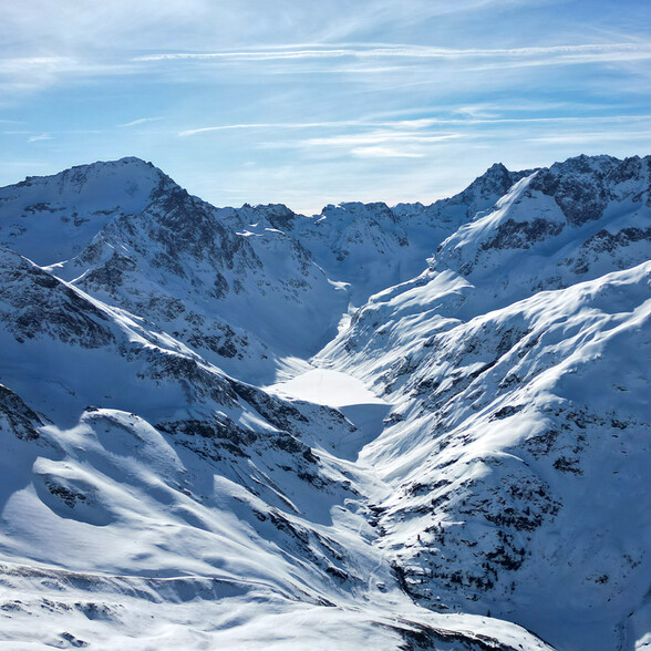 Blick auf ein verschneites Hochtal mit dem gefrorenen Kartellsee, umgeben von schroffen, weißen Gipfeln unter blauem Himmel.