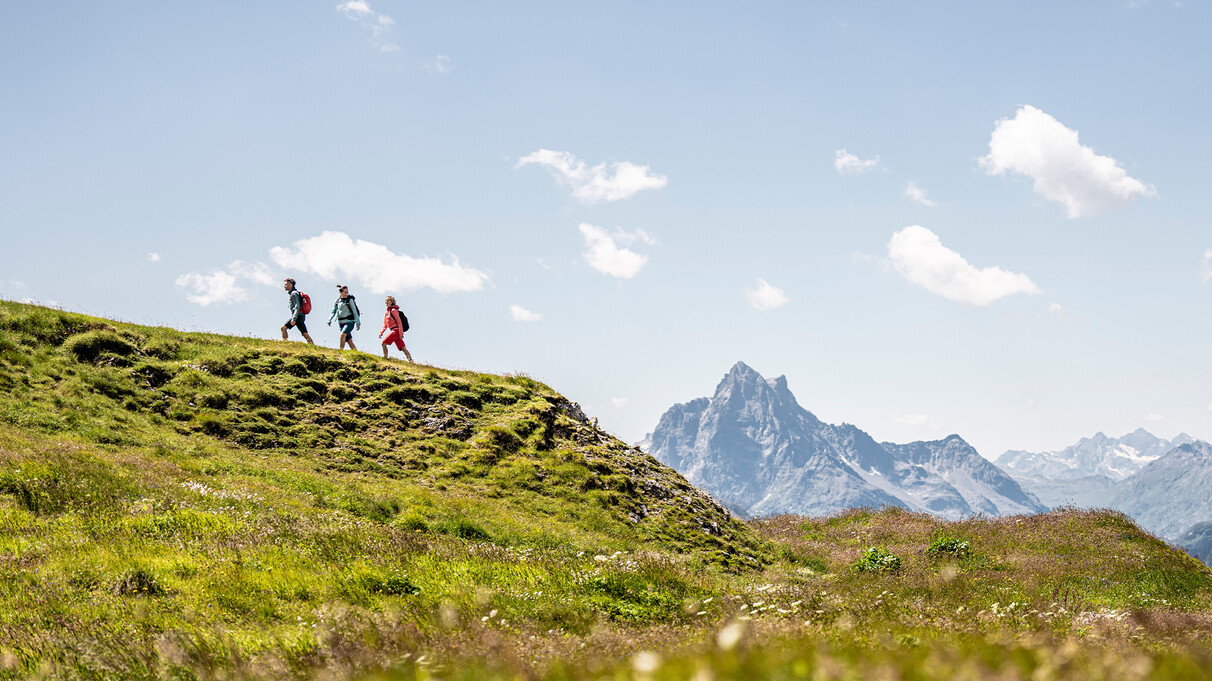 Three hikers walk along a grassy ridge above St. Anton am Arlberg, enjoying a wide view of the majestic alpine landscape. In the background rises the striking peak of the Parseierspitze.