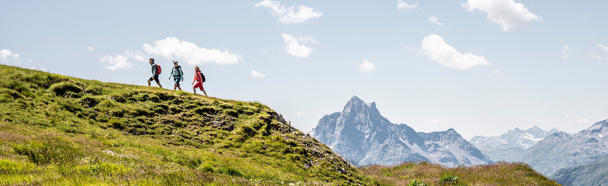 Drei Wandernde gehen auf einem grasbewachsenen Höhenrücken oberhalb von St. Anton am Arlberg mit freiem Blick auf die majestätische Berglandschaft. Im Hintergrund ragt der markante Gipfel der Parseierspitze empor.