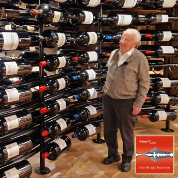 Adi Werners steht lächelnd in einem stilvoll beleuchteten Weinkeller vor einem Wandregal mit vielen liegenden Weinflaschen in Magnumgröße.