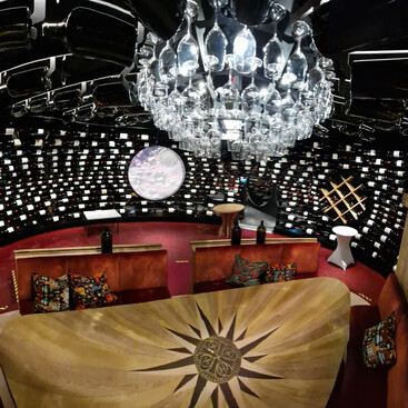 Panoramablick auf einen runden Weinkeller mit rotem Teppich, verzierten Sitznischen, großen Weinflaschen und einer Decke aus hängenden Weingläsern unter einem runden Kronleuchter.