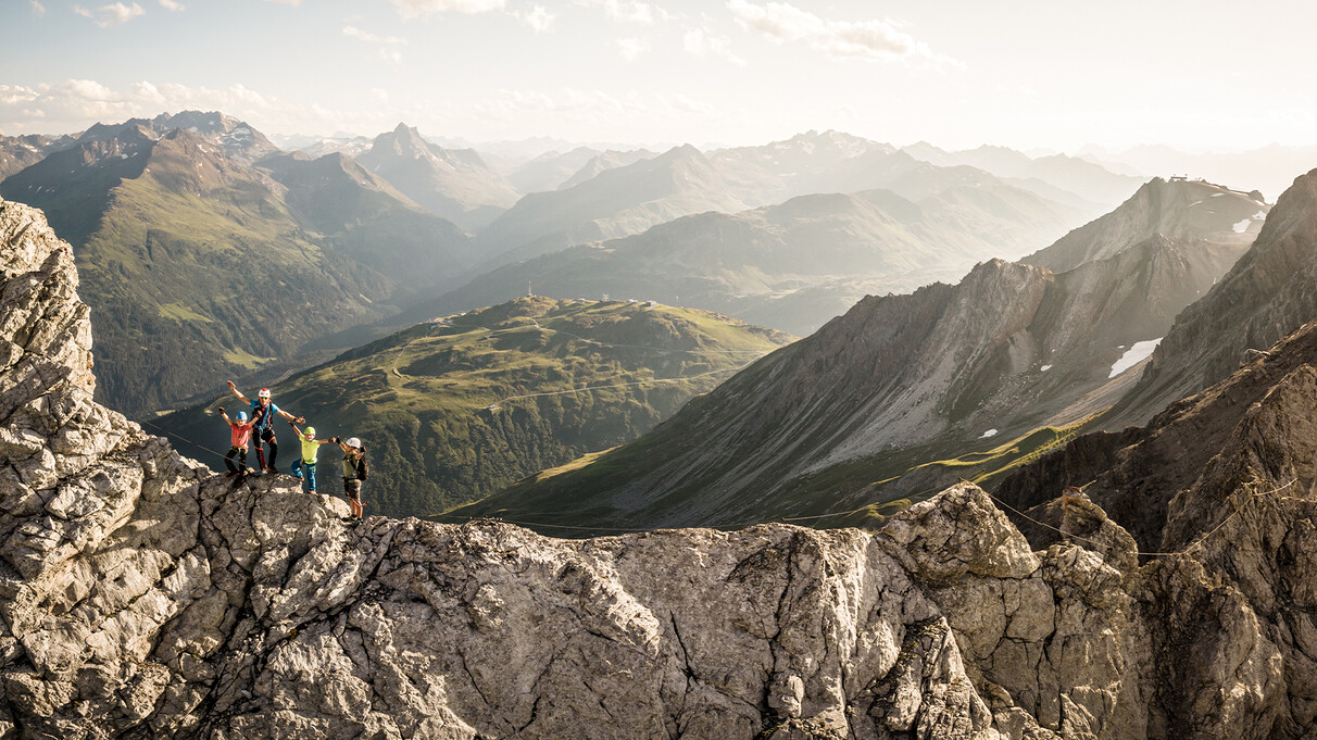 A group climbs a rocky ridge over St. Anton am Arlberg with safety gear, overlooking sunlit alpine valleys and rugged mountain peaks.