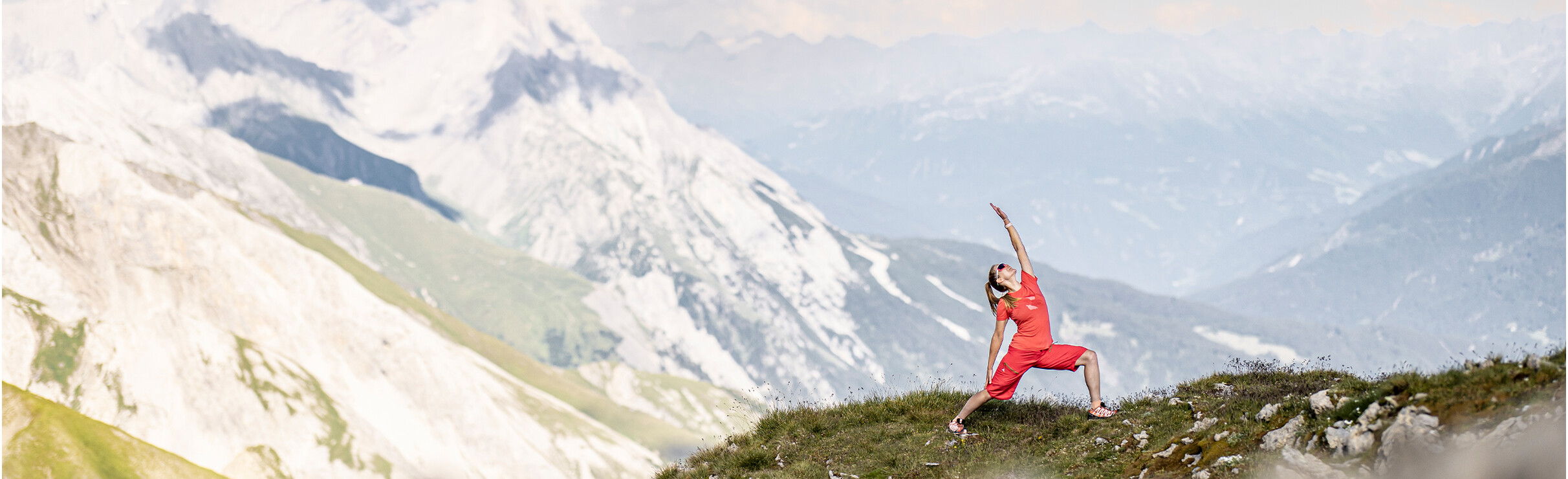 Eine Frau macht eine Yogapose auf einer Almwiese mit Ausblick auf verschneite Berge unter klarem Himmel.