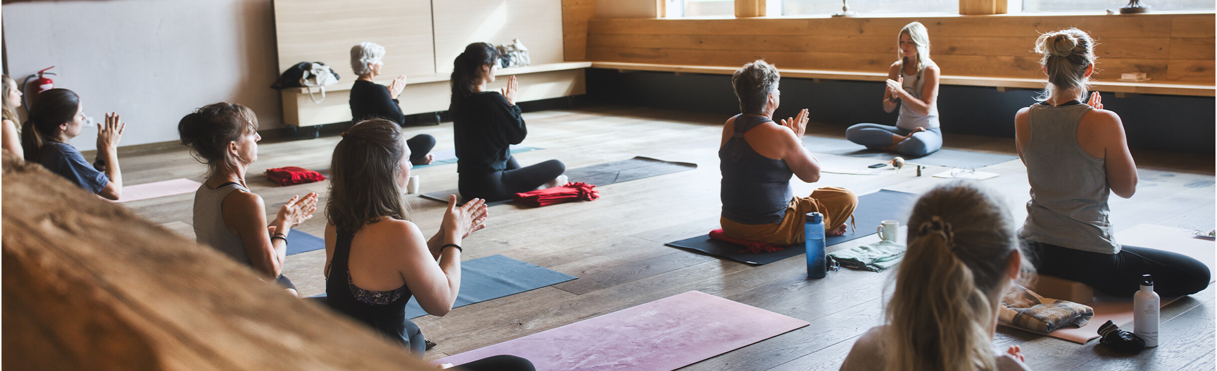 Mehrere Personen sitzen auf Yogamatten in einem Holzstudio und meditieren gemeinsam im Schneidersitz.