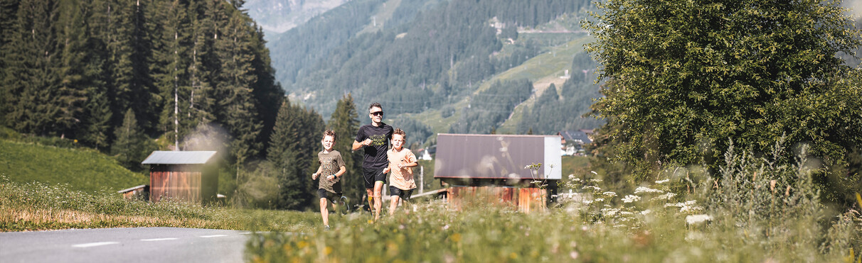 Vater läuft mit zwei Kindern durch blühende Wiesen vor einer idyllischen Bergkulisse.