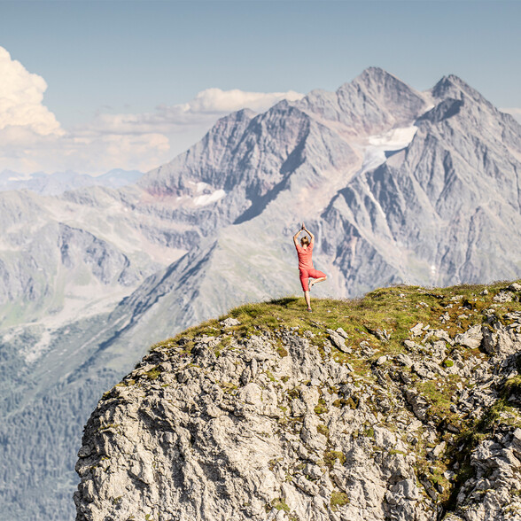 Eine Frau steht im Baum auf einer Bergwiese mit eindrucksvoller Aussicht auf schroffe Gipfel.