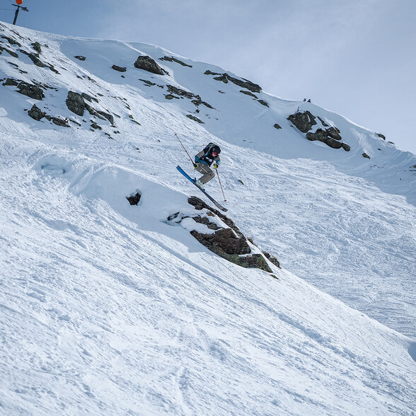 Ein Freeride-Skifahrer springt dynamisch über eine Felskante im verschneiten Gelände von St. Anton am Arlberg. Der Himmel ist leicht bewölkt und die winterliche Berglandschaft umgibt ihn.